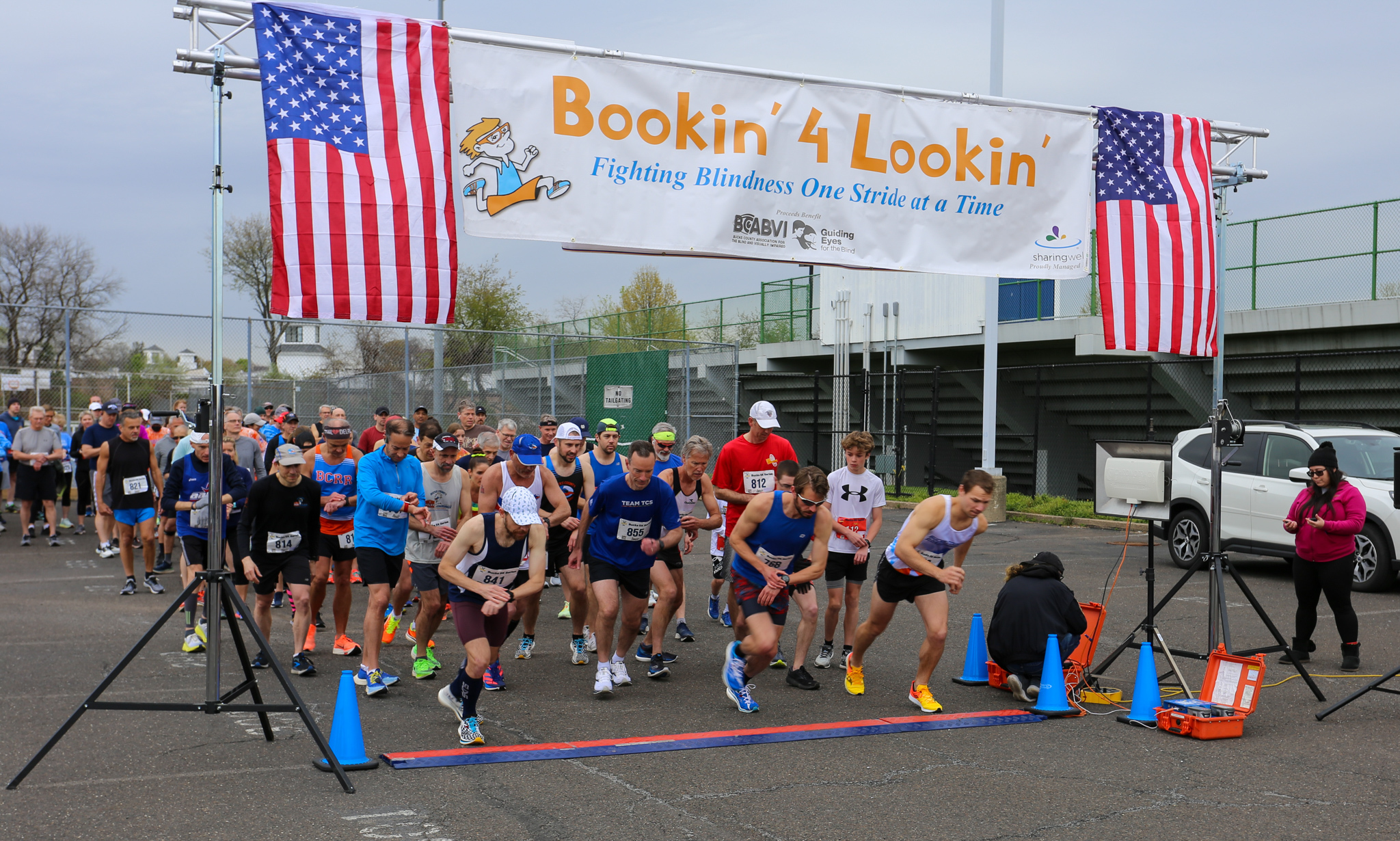 Runners surging off the start line beneath the Bookin' for Lookin' Race For Sight banner, flanked by American flags
